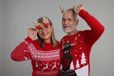Photo of Senior couple in Christmas sweaters, reindeer headband and funny glasses on grey background