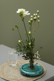 Stylish ikebana with beautiful flowers, green branches and glass of water on gray table near olive wall