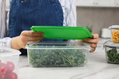 Woman sealing glass container with greens at white marble table in kitchen, closeup