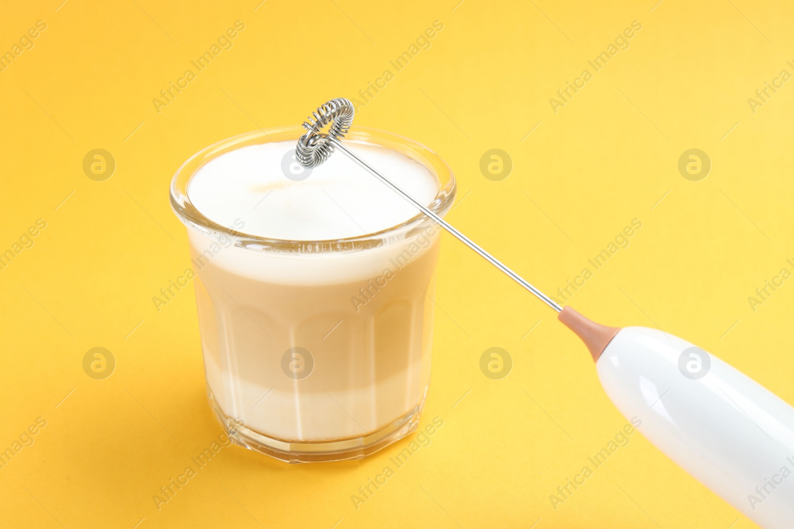 Photo of Mini mixer (milk frother) and tasty cappuccino in glass on yellow background