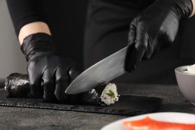 Photo of Chef in gloves cutting sushi roll at dark textured table, closeup
