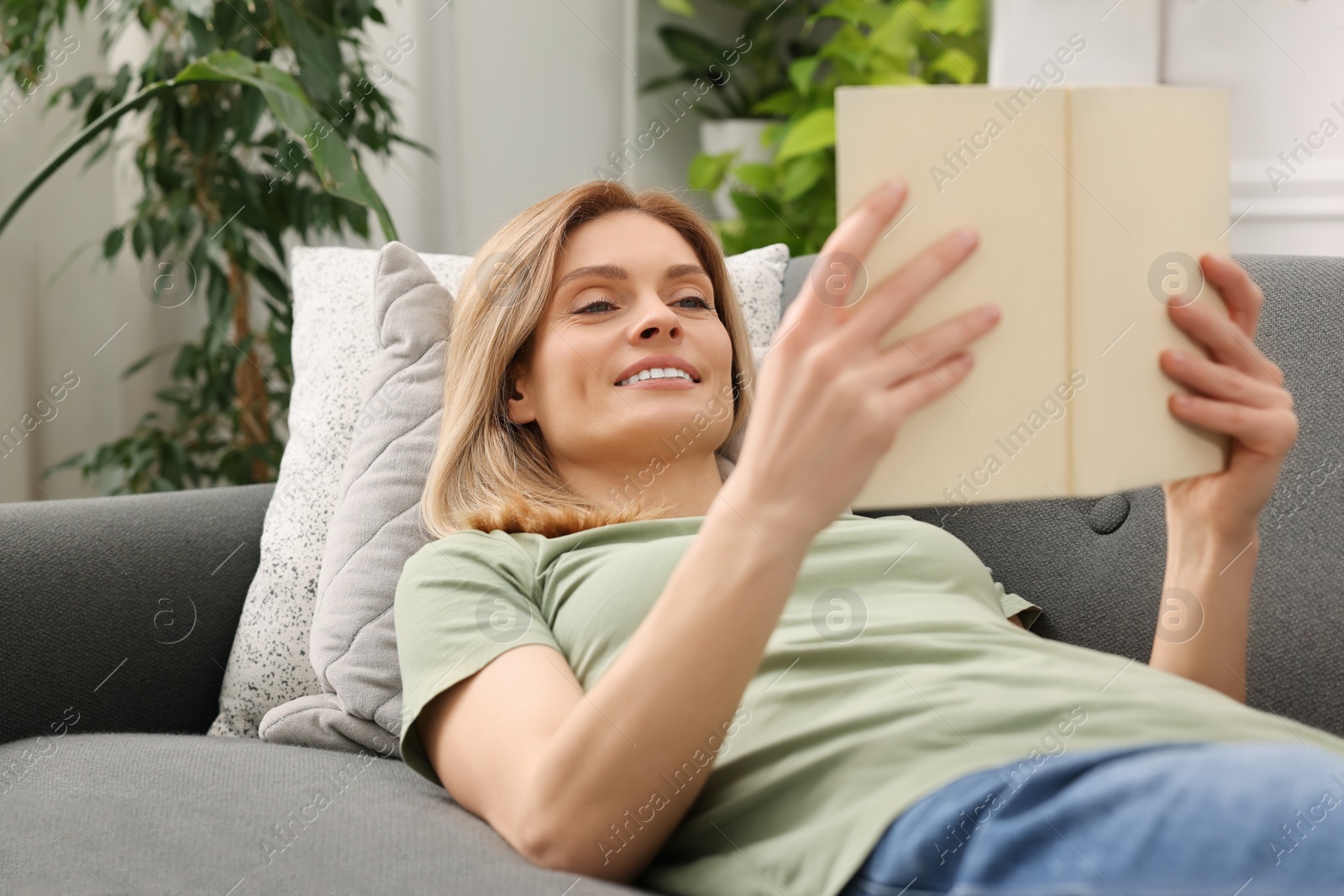 Photo of Woman reading book on sofa in room with houseplants