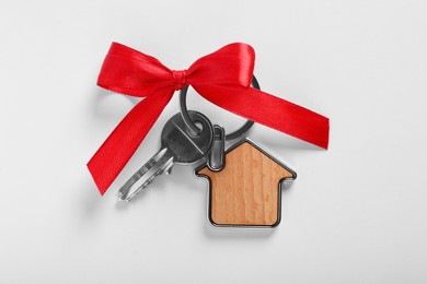 Key with trinket in shape of house and red bow on light grey background, top view. Housewarming party