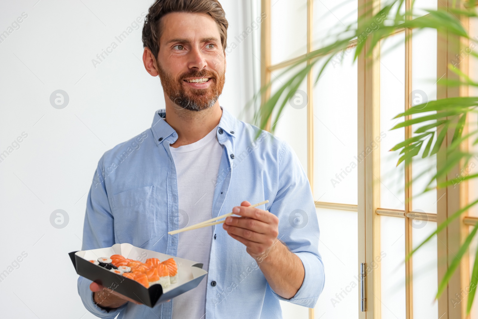 Photo of Happy man eating sushi rolls with chopsticks indoors