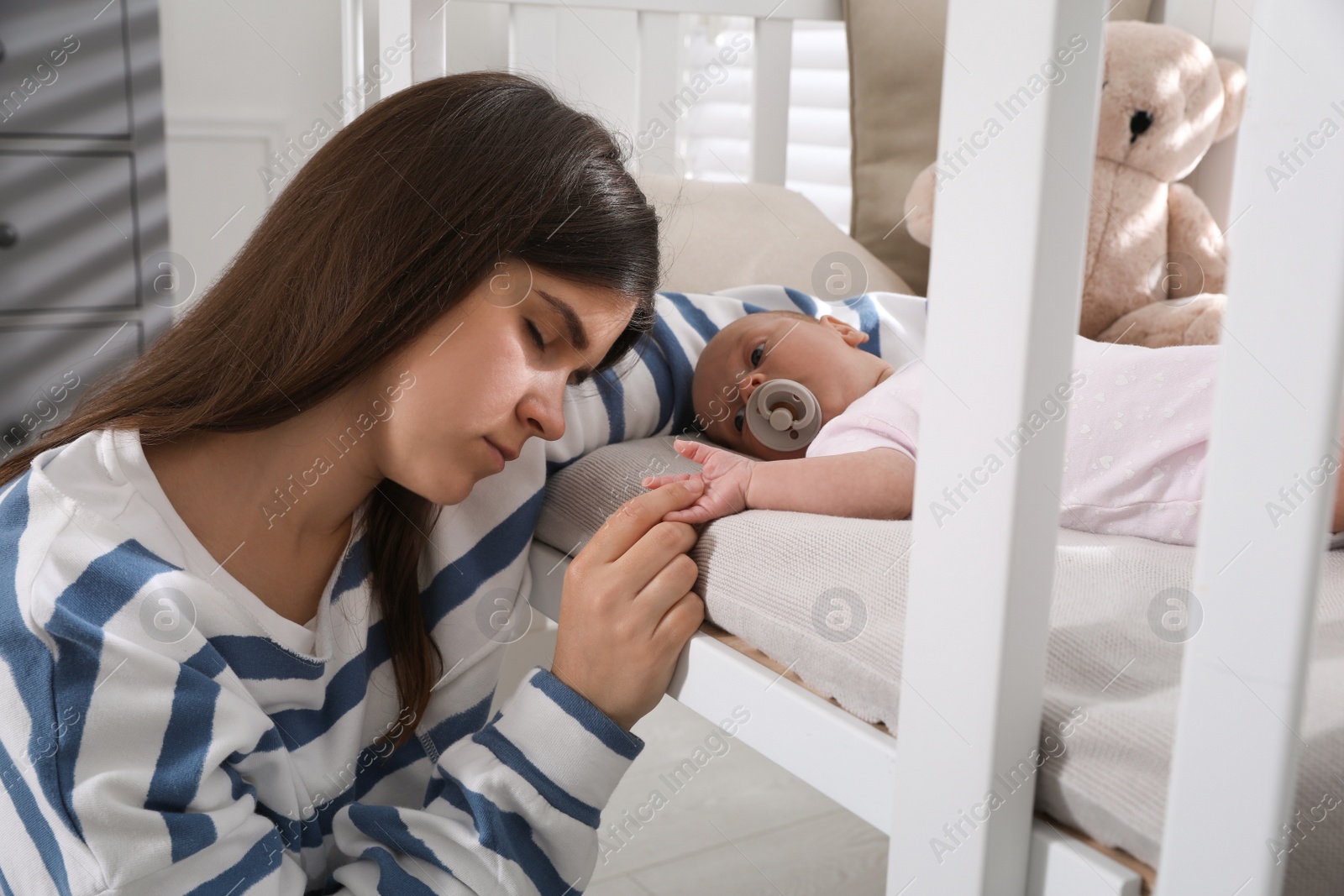 Photo of Tired young mother sleeping near open crib with her baby in children's room