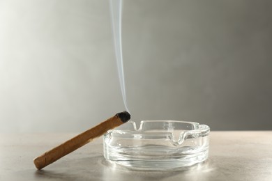Photo of Smoldering cigar near glass ashtray on light grey table, space for text
