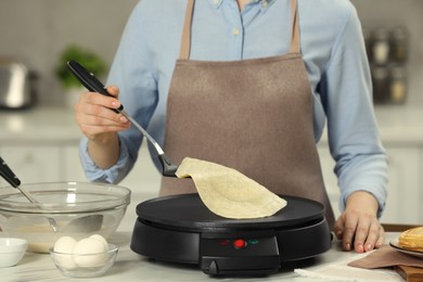 Woman cooking delicious crepe on electric maker at white marble table in kitchen, closeup