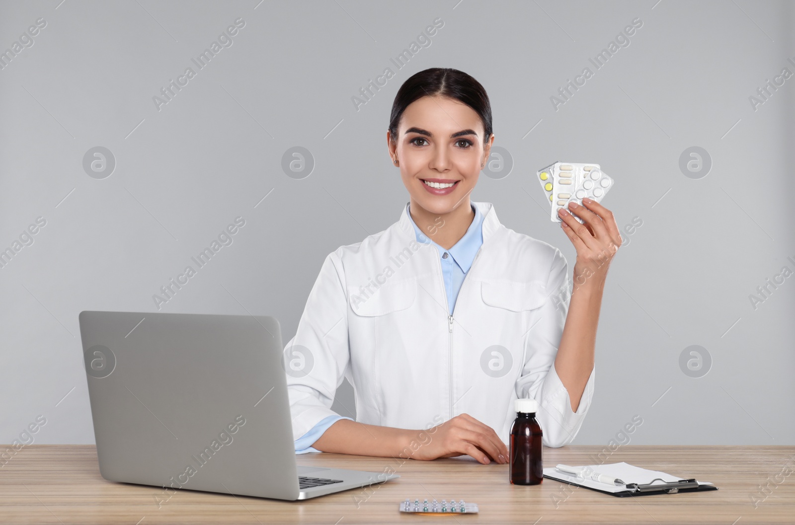 Photo of Professional pharmacist with pills and laptop at table against light grey background