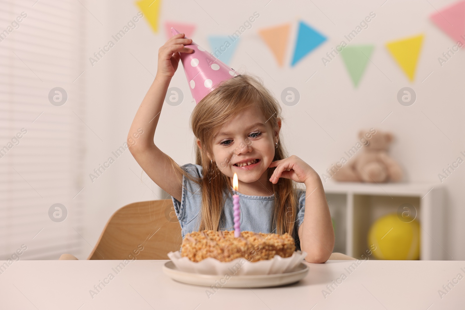 Photo of Cute girl in party hat with birthday cake at table indoors