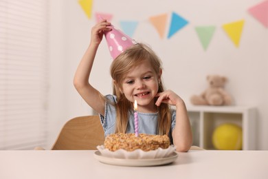 Cute girl in party hat with birthday cake at table indoors