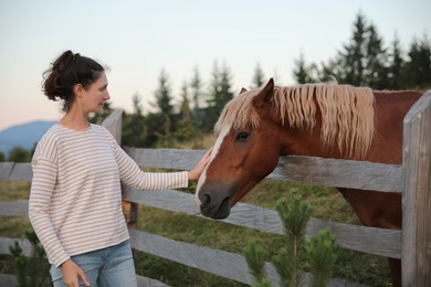 Photo of Woman stroking beautiful horse near wooden fence outdoors. Lovely domesticated pet