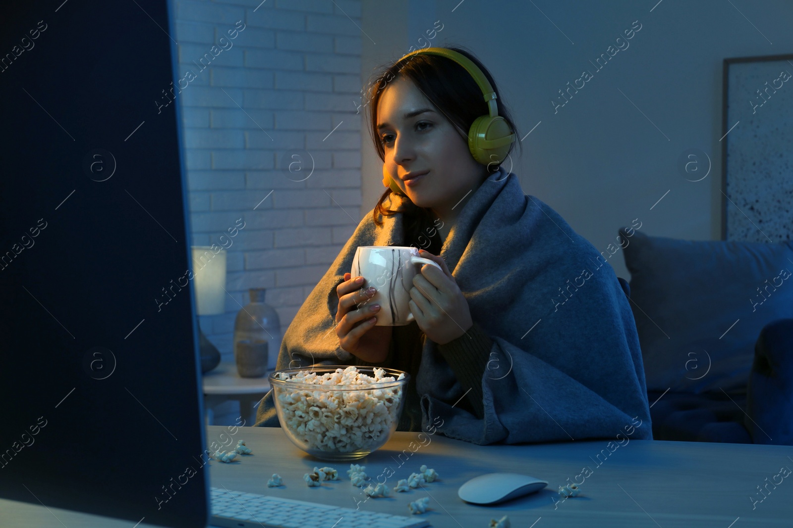 Photo of Beautiful young woman under blanket in headphones with popcorn and cup of drink watching film on computer at table indoors at night