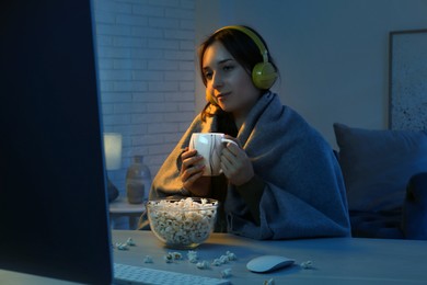 Beautiful young woman under blanket in headphones with popcorn and cup of drink watching film on computer at table indoors at night