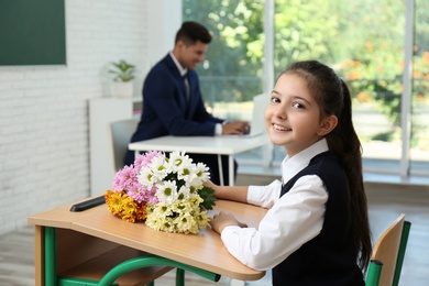 Happy schoolgirl with bouquet sitting at desk in classroom. Teacher's day