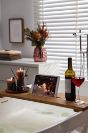 Wooden tray with tablet, wine and burning candles on bathtub in bathroom