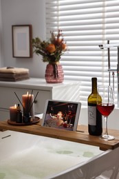 Photo of Wooden tray with tablet, wine and burning candles on bathtub in bathroom