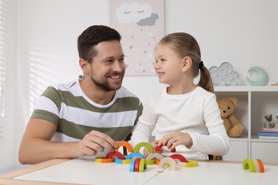 Motor skills development. Father helping his daughter to play with colorful wooden arcs at white table in room