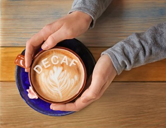 Woman holding cup of aromatic decaf coffee with foam at wooden table, top view