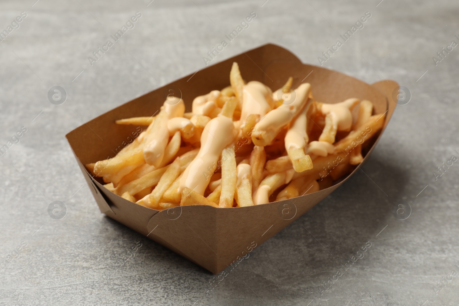 Photo of Tasty potato fries and cheese sauce in paper container on grey table, closeup