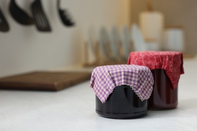 Jars of jams covered with beeswax food wrap on white table indoors, space for text