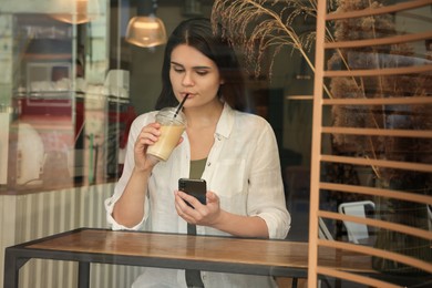 Photo of Young beautiful woman with smartphone drinking coffee in cafe, view through window glass