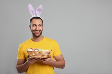 Happy African American man in bunny ears headband holding wicker tray with Easter eggs on gray background, space for text