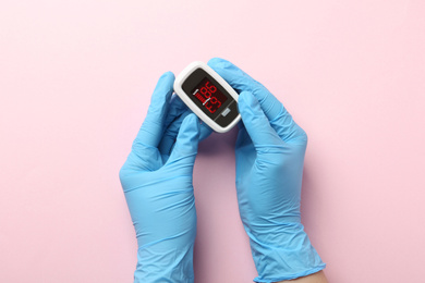 Photo of Doctor in gloves holding fingertip pulse oximeter on pink background, top view