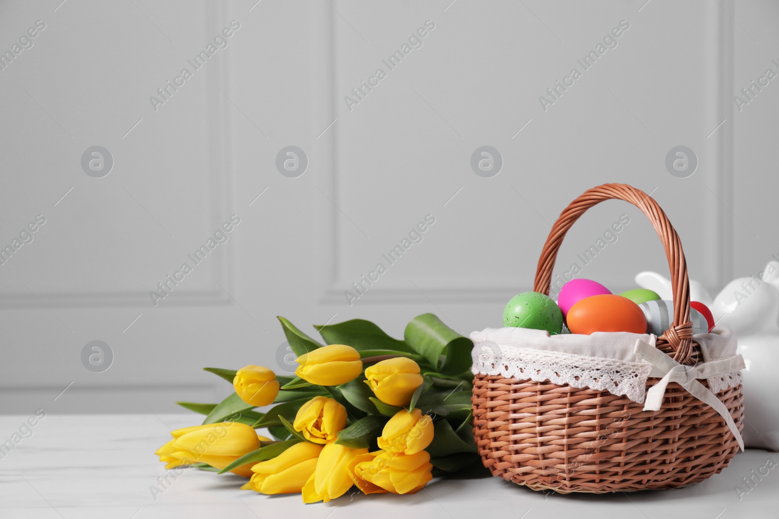 Photo of Easter basket with painted eggs near bouquet of tulips on white marble table. Space for text