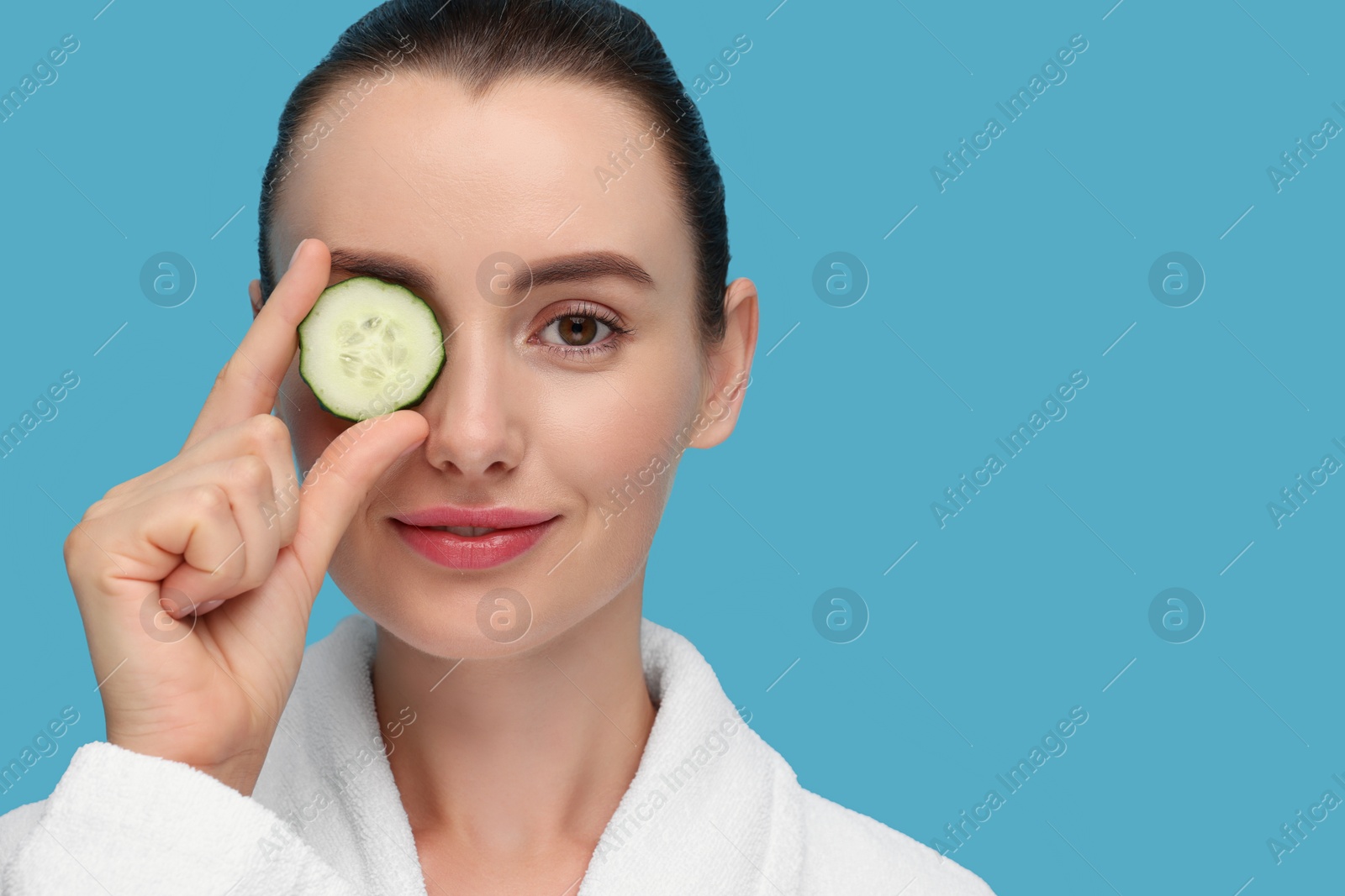 Photo of Beautiful woman covering eye with piece of cucumber on light blue background. Space for text