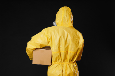 Photo of Man wearing chemical protective suit with cardboard box on black background, back view. Prevention of virus spread