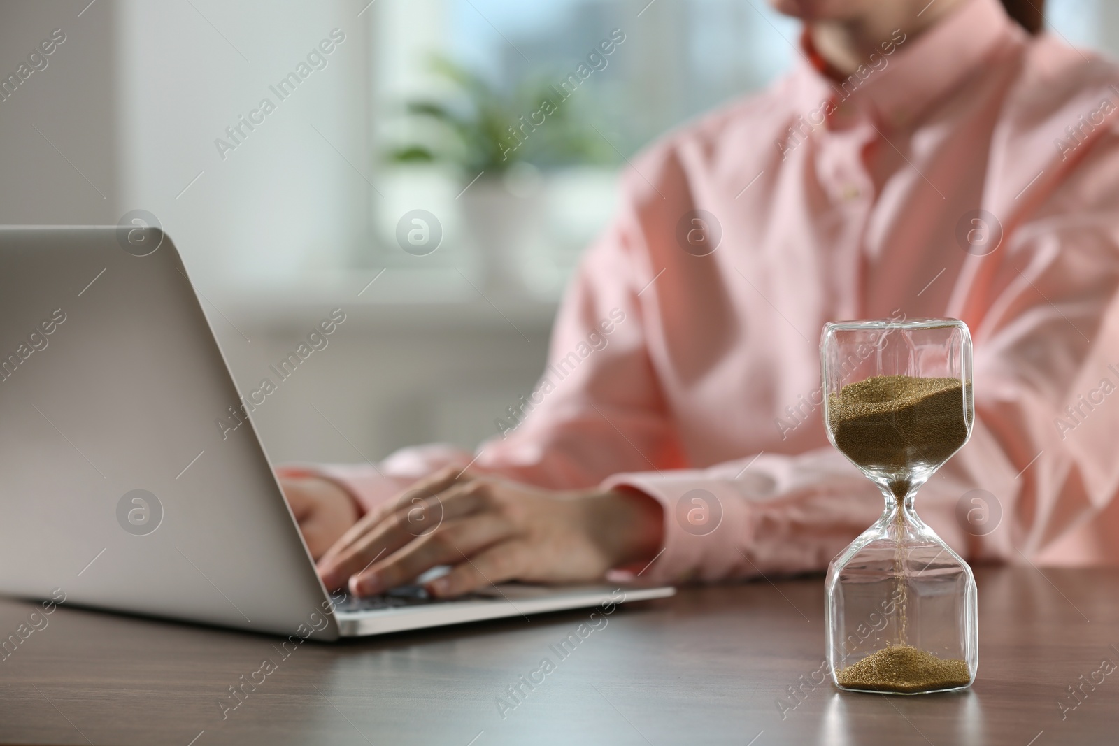 Photo of Hourglass with flowing sand on wooden table, selective focus. Man using laptop indoors