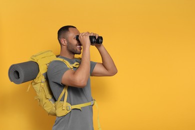 Photo of Happy tourist with backpack looking through binoculars on yellow background, space for text