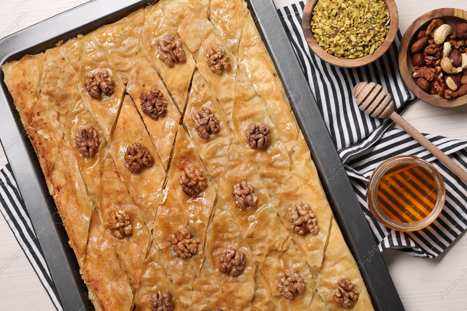 Photo of Delicious baklava with walnuts in baking pan, honey and nuts on white wooden table, flat lay