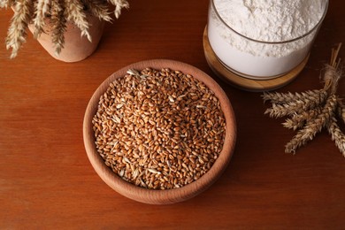 Photo of Wheat grains in bowl, spikes and flour on wooden table, above view