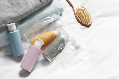 Photo of Cosmetic travel kit. Plastic bag with small containers of personal care products, brush and stack of clothes on bed