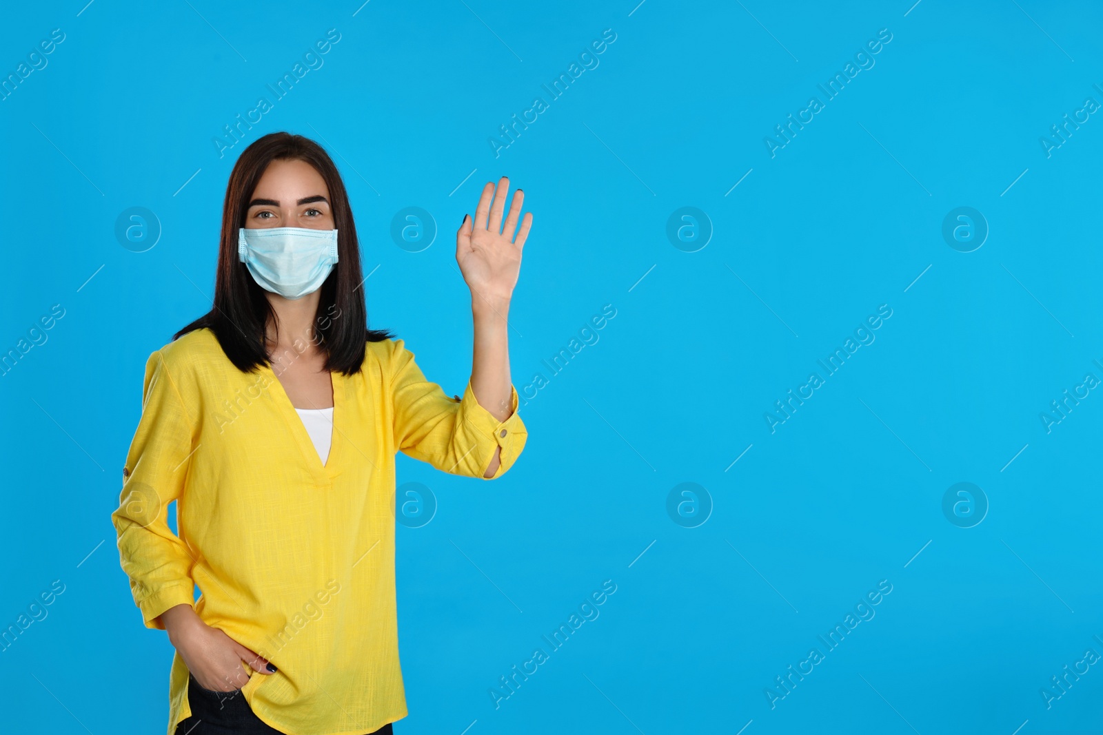 Photo of Young woman in protective mask showing hello gesture on light blue background, space for text. Keeping social distance during coronavirus pandemic