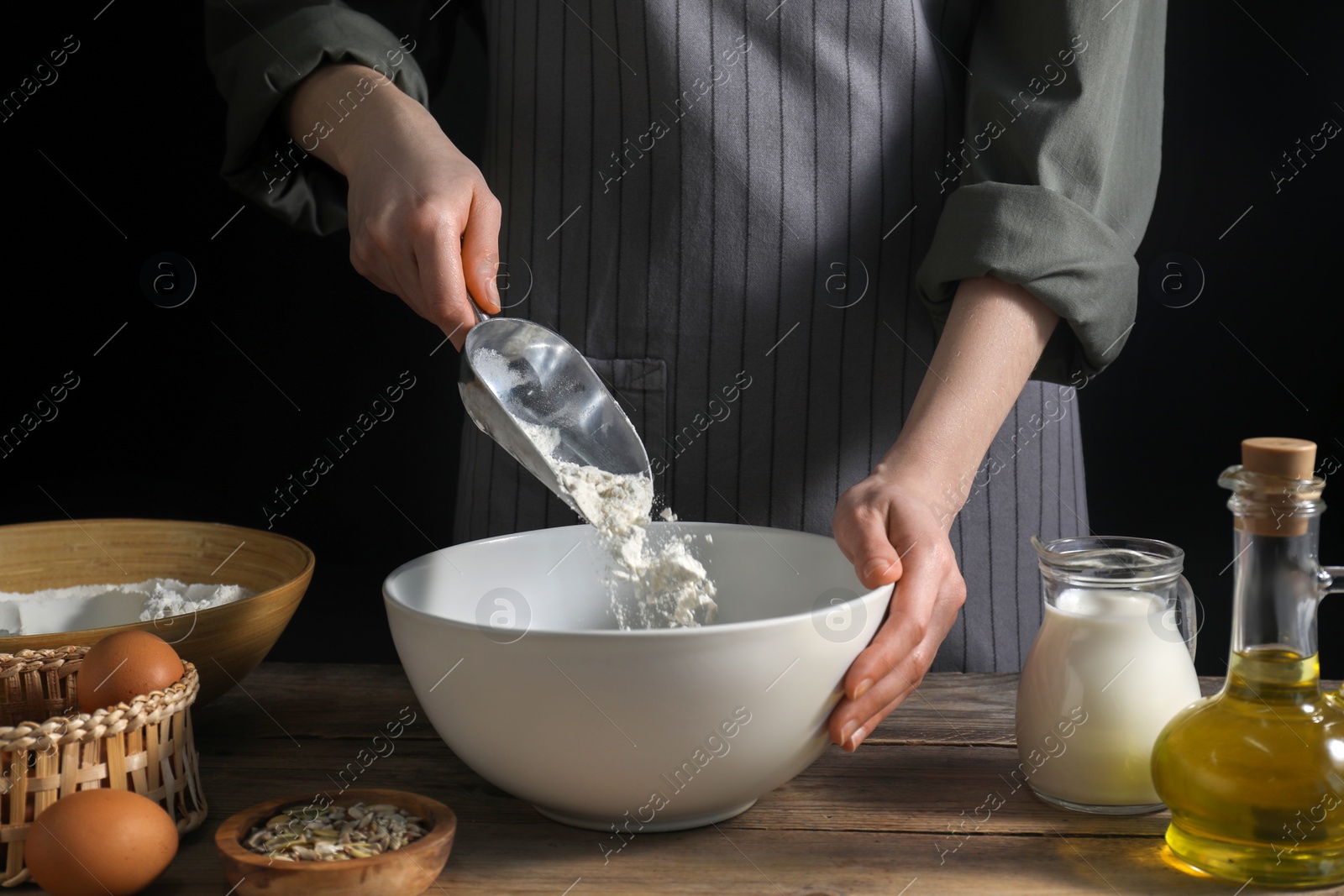 Photo of Making bread. Woman adding flour into bowl at wooden table on dark background, closeup