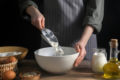 Making bread. Woman adding flour into bowl at wooden table on dark background, closeup