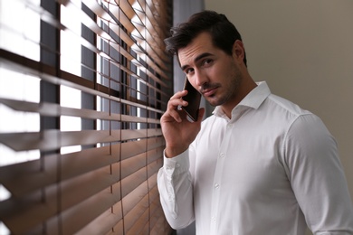 Photo of Handsome young man talking on smartphone near window indoors