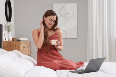 Happy woman with cup of drink near laptop on bed in bedroom