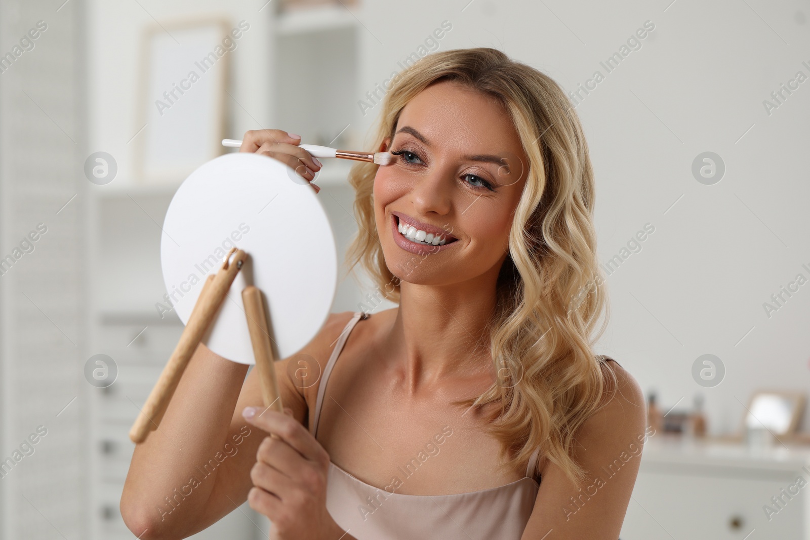 Photo of Beautiful makeup. Smiling woman applying eyeshadows in front of mirror indoors