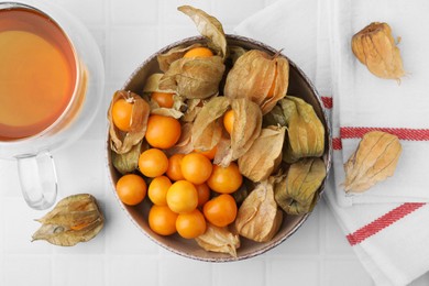 Ripe physalis fruits with calyxes in bowl and cup of tea on white tiled table, flat lay