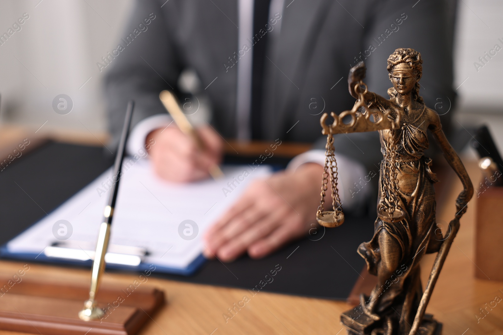 Photo of Notary writing notes at wooden table in office, focus on statue of Lady Justice