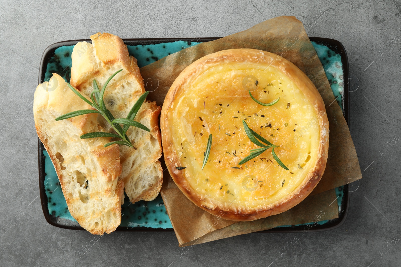 Photo of Plate with tasty baked brie cheese, bread and rosemary on grey table, top view