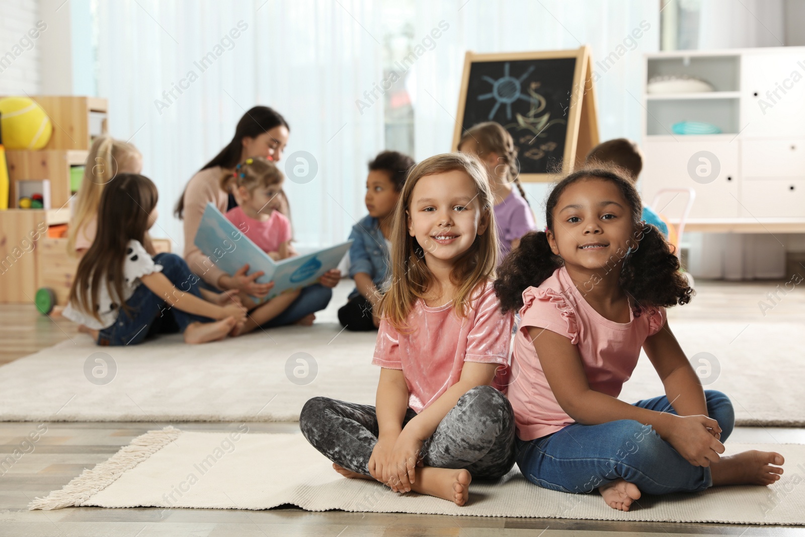 Photo of Cute girls sitting on floor while kindergarten teacher reading book to other children indoors