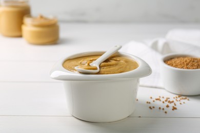 Photo of Spoon and bowl of tasty mustard sauce on white wooden table, closeup