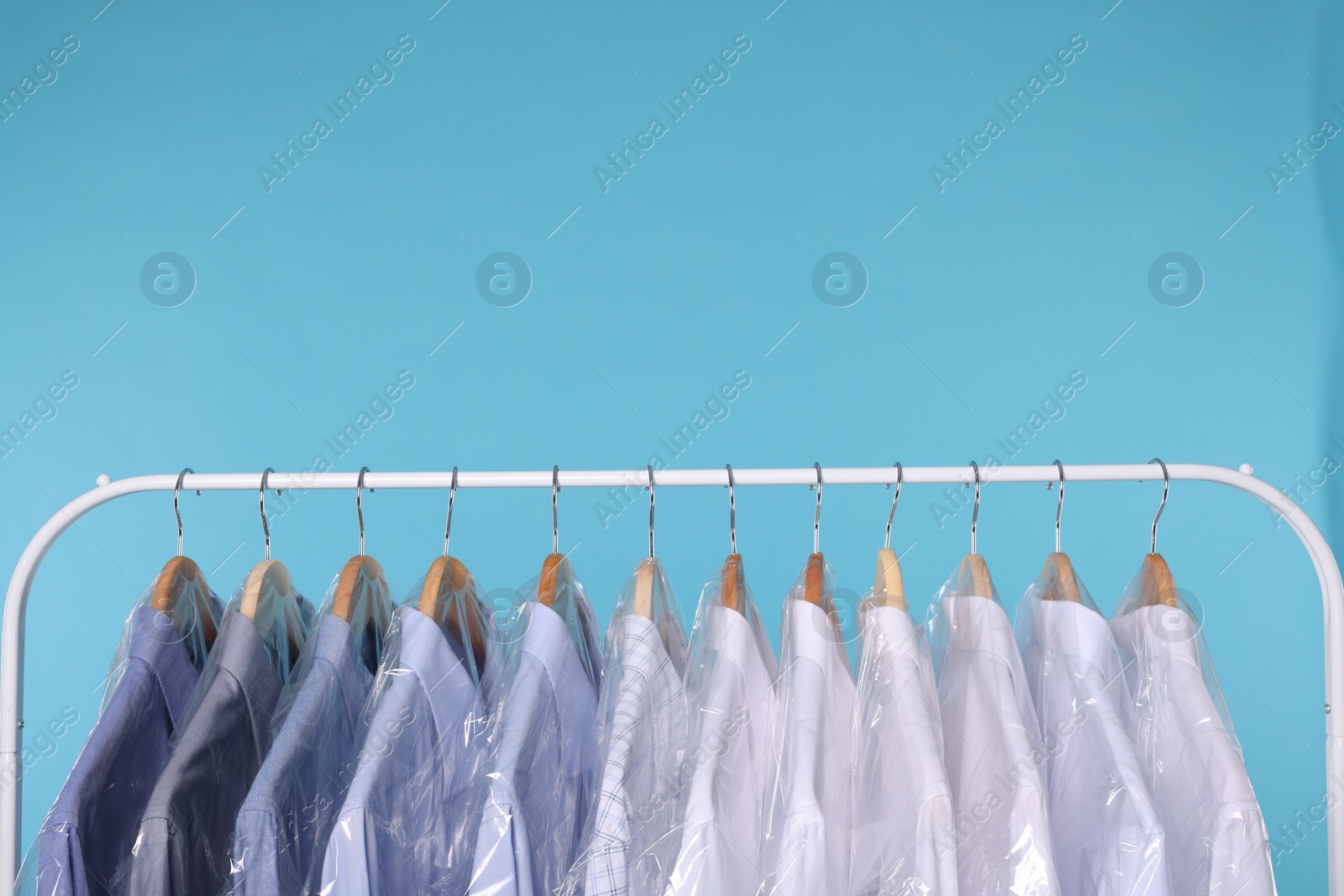 Photo of Dry-cleaning service. Many different clothes in plastic bags hanging on rack against light blue background