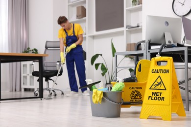Photo of Cleaning service worker washing floor with mop, focus on bucket with supplies and wet floor sign in office