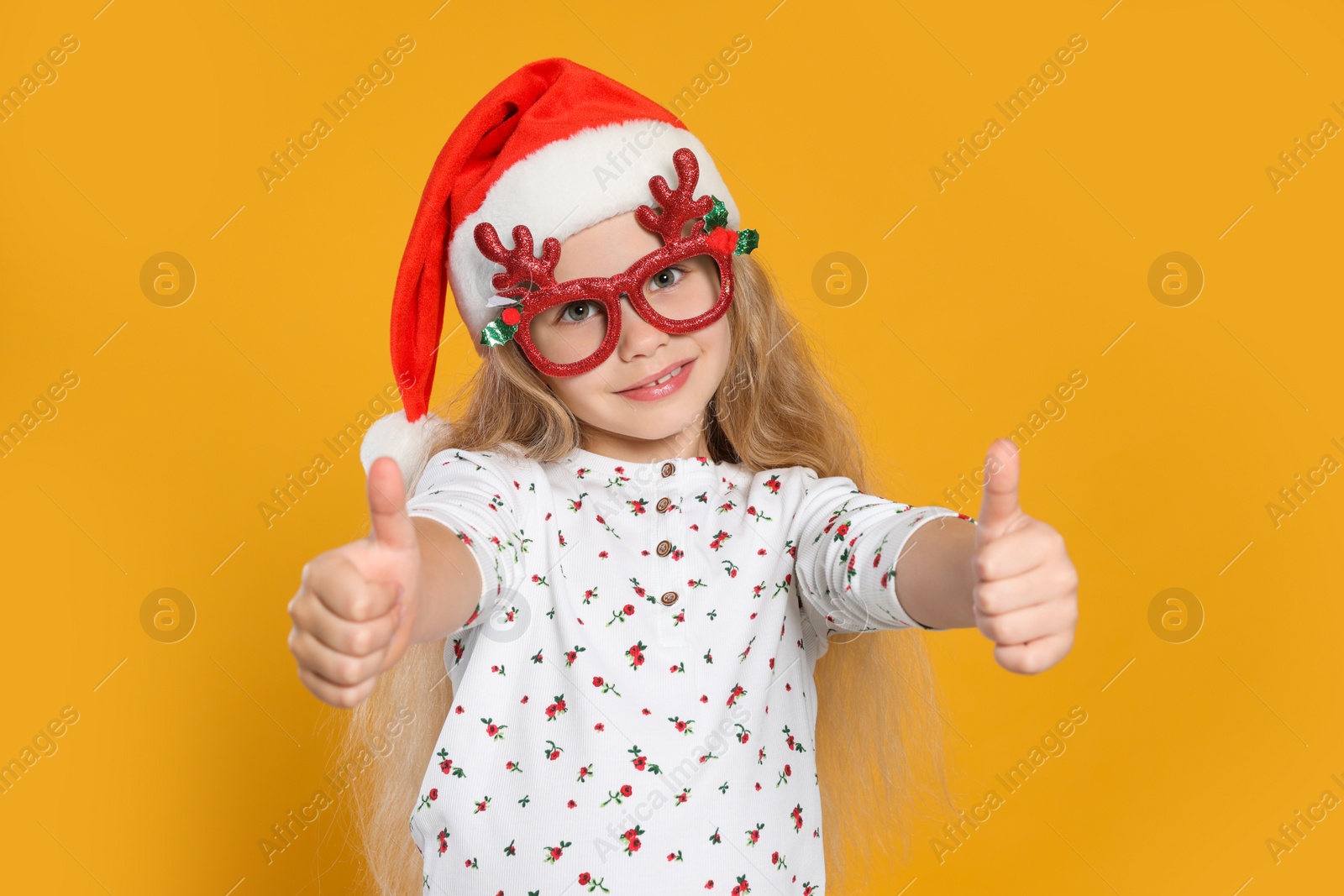 Photo of Girl in decorative eyeglasses and Santa hat showing thumbs up on orange background