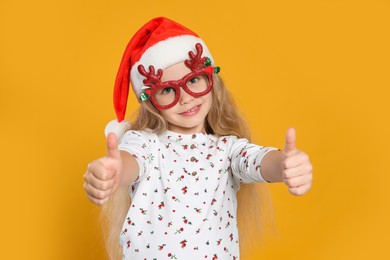 Girl in decorative eyeglasses and Santa hat showing thumbs up on orange background
